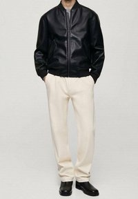 Homme portant une veste bomber en cuir noir, un pantalon couleur crème et des bottes noires, debout les mains dans les poches devant un fond uni.