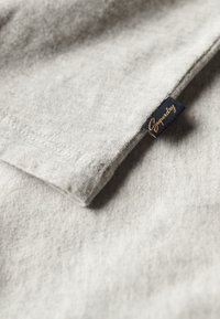 Close-up van een lichtgrijze stoffen mouw met een klein zwart label met in gele krullende letters bedrukt, genaaid in de naad.