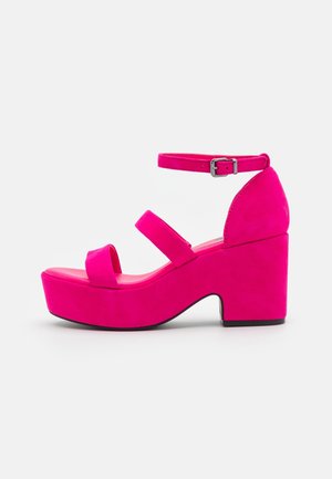 Jasnoróżowe sandały na platformie z dwoma paskami z przodu, masywnym słupkiem oraz regulowanym paskiem na kostkę na białym tle.