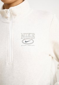 Lys kremfarget fleece-pullover med halvdelt glidelås i kragen. Har en Nike-logo og tekst i mørkegrått. Myk tekstur, varm og sporty design.
