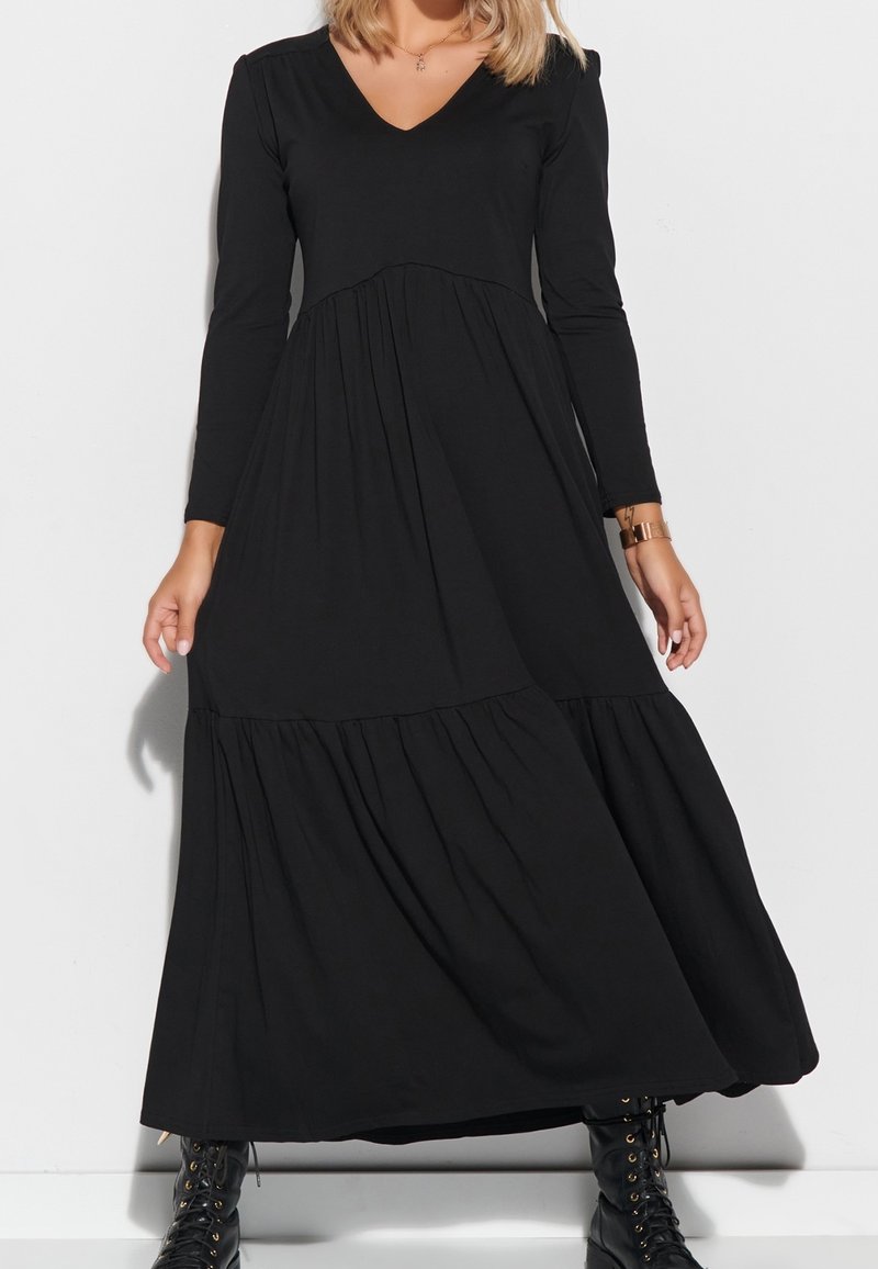 Femme portant une longue robe noire à manches longues et col en V, associée à des bottes noires lacées et une montre au poignet gauche.