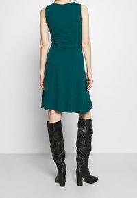 Robe sans manches couleur sarcelle avec une jupe évasée, en tissu lisse. Associée à des bottes en cuir noir souple montant jusqu'aux genoux avec des talons blocs.