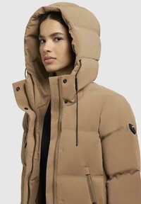 Beige puffjakke med høj hætte, lynlås og trykknaplukninger, elastisk snøre og sidelommer med lynlås; har et quiltet, broderet design.