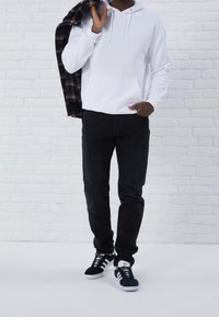 Sudadera blanca con bolsillo delantero, jeans negros y zapatillas negras con detalles en blanco, sosteniendo una camisa de cuadros contra un fondo de ladrillo blanco.