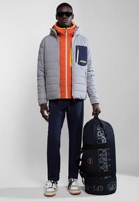 Chaqueta gris acolchada con una capa interior naranja, bolsillo azul marino. Pantalones azul marino y bolsa de deporte azul marino con estampado. Modelo luciendo gafas de sol y zapatillas.