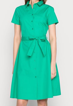 Frau trägt ein knielanges, kurzärmeliges grünes Hemdkleid mit einem gebundenen Gürtel in der Taille.