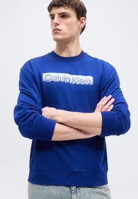 Ung mand med armene krydset iført en blå sweatshirt med "Calvin Klein"-logo, lyse jeans, kigger til venstre mod en ensartet lys baggrund.