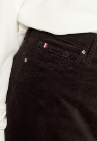 Väriharmaat corduroy-housut, joissa on teksturoitu pinta, ja joissa on pieni punainen, valkoinen ja sininen logo taskussa sekä kultaiset yksityiskohdat.