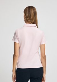 Camisa polo de manga corta en rosa claro, textura suave, cuello clásico, diseño entallado, con un dobladillo recto y sin patrones ni detalles visibles.