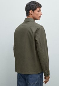 Oliven grøn, letvægtsjakke med krave, knaplukning ved ærmerne og lige skæring, parret med blå denimjeans.