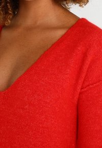 Pull en tricot rouge, doux, avec un décolleté en V profond et des manches longues. Le tissu est texturé, avec des fibres visibles et une coupe décontractée.