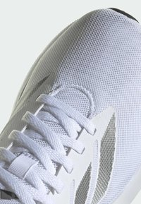 Baskets blanches avec un dessus en maille, lacets plats et accents argentés. Présente un bout arrondi et des détails texturés sur le côté.