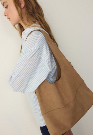 Frau mit langen, hellbraunen Haaren, die ein blau-weiß gestreiftes Hemd trägt und eine große, hellbraune Wildleder-Schultertasche vor einer schlichten Wand trägt.