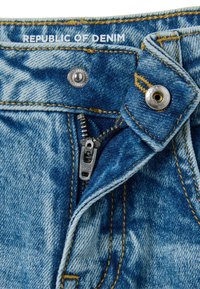 Jeans de mezclilla azul con una cremallera y cierre de botón visibles. Presenta costuras en contraste, un ligero desgastado y una etiqueta de marca que dice "REPÚBLICA DE DENIM".