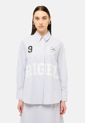 Femme aux cheveux raides et foncés portant une chemise blanche et bleue à fines rayures, manches longues, avec le numéro 9 et des écussons de logo sur la poitrine.