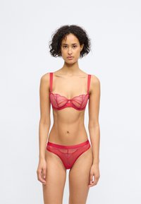 Conjunto de lingerie vermelho com painéis de malha transparente, soutien com aro e costura decorativa, e cuecas combinando com um padrão intricado.