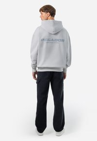 Grå hættetrøje med "PEGADOR" trykt på ryggen, kombineret med mørke cargo-bukser og hvide sneakers. Løs pasform og blød tekstur.