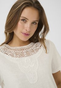T-shirt bianco a maniche corte con dettagli in pizzo intricato attorno al collo, caratterizzato da motivi geometrici e accenti testurizzati.