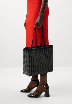 Zwarte geometrische totebag met een patroonachtige textuur, dubbele schouderbanden en een gestructureerde vorm, gecombineerd met glanzende zwarte schoenen met hakken.