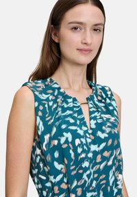 Türkiz blúz kék és barack színű mintázattal, lekerekített nyakkivágással, redőzött részletekkel és egy minimális fém díszítéssel a nyakkivágásnál.