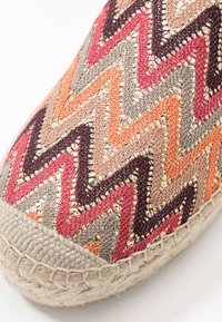 Närbild av en skotå med vävda zigzagmönster i orange, röd, rosa, beige och brun, med en flätad sula av naturliga fibrer.