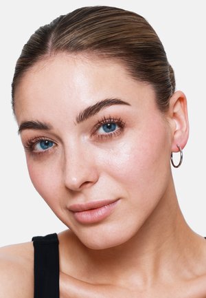 Nahaufnahme einer Person mit heller Haut, glatter Textur, blauen Augen und minimalem Make-up. Sie trägt kleine silberne Creolen und hat die Haare nach hinten frisiert.