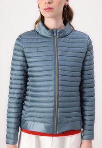 Lys blå pufferjakke med vertikal quiltet design, frontlynlås, stående krave og glat tekstur. Bæres over en rød bluse.