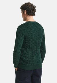 Dunkelgrüner Kabelstrickpullover mit gerippten Bündchen und Saum, einem strukturierten Muster, von hinten mit taillierter Passform gezeigt.