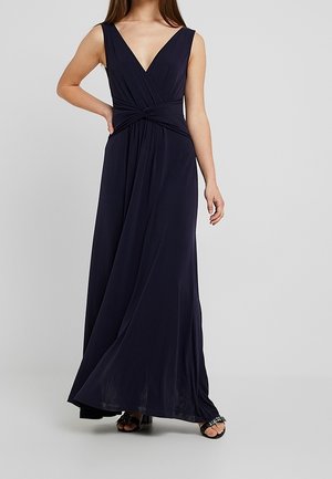 Kobieta w granatowej, bezrękawowej sukni maxi z głębokim dekoltem w szpic i skręconym detalem w talii, w połączeniu z czarnymi sandałami na otwartym palcu.