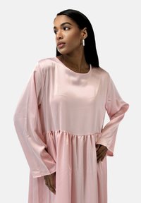 Robe en satin rose pâle avec manches longues et taille froncée. Texture lisse et encolure ronde, créant une silhouette décontractée.