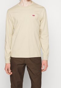 Beige Langarm-T-Shirt aus Baumwolle, mit einem kleinen roten Levi's-Logo auf der linken Brust, Rundhalsausschnitt und gerippten Bündchen.