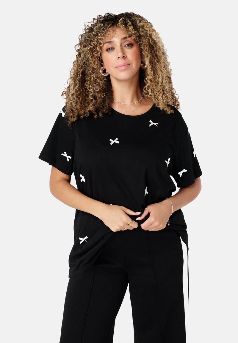 Zwarte T-shirt met korte mouwen van zacht materiaal, voorzien van witte strikborduursels verspreid over de voorkant. Losse pasvorm met ronde halslijn.