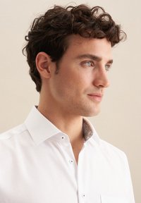 Biała koszula z wykrojem kołnierza, ozdobiona wzorzystą podszewką i minimalnym detalem guzika. Gładka bawełniana tekstura.