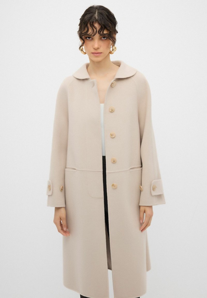 Cappotto in lana beige con ampio colletto, bottoni frontali e tasche laterali. Presenta maniche lunghe con polsini abbottonati e una finitura testurizzata.