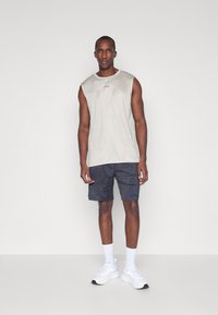 Ärmelloses beiges Sportshirt mit geometrischem Muster, kombiniert mit dunkel gemusterten Shorts und weißen Sneakers mit Knöchelsocken.
