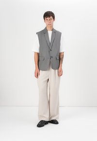 Šedý bezrukávový blazer se dvěma knoflíky, kombinovaný s bílým tričkem a volnými béžovými kalhotami. Model stojí na neutrálním pozadí.