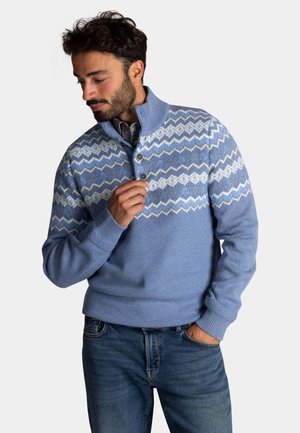 Lichtblauwe gebreide trui met een hoge kraag, voorzien van een zigzagpatroon in wit en donkerder blauw, met een drieknopensdetail bij de hals.