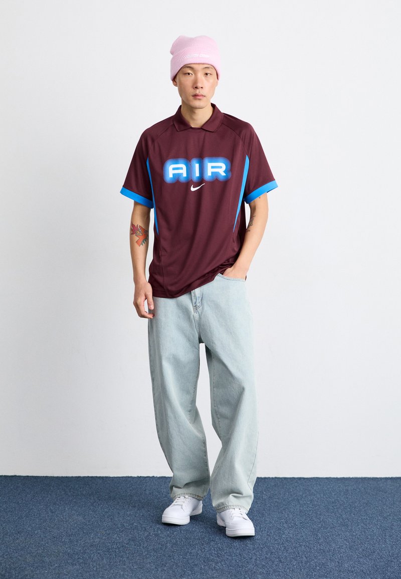 Polo shirt bordeaux avec des accents bleus, arborant le mot "AIR" en texte gras et un logo Nike. Associé à un jean baggy bleu clair et des baskets blanches.