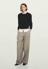 Maglione nero a maglia, camicia bianca con colletto e pantaloni plissettati grigi chiari. Il modello è in piedi con le mani in tasca, indossando scarpe nere.