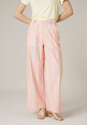 Une personne porte un pantalon ample taille haute rose avec des rayures verticales multicolores et des sandales à talons blocs dorés à brides, debout les mains derrière le dos.