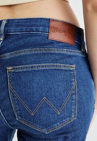 Blue denim jeans with a textured finish, featuring a brown leather patch embossed with "Wrangler" and a zigzag stitch pattern on the back pockets.