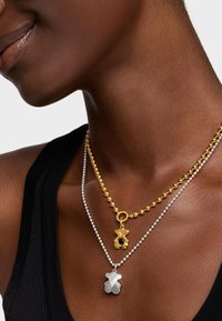 Collane in oro e argento sovrapposte, con un pendente in oro con texture e un pendente in argento liscio a forma di orso.