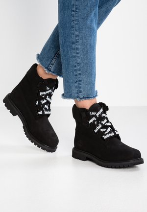 Pieds portant des bottes Timberland noires avec des lacets blancs portant le logo et un jean bleu à ourlet effiloché, debout sur un sol blanc.