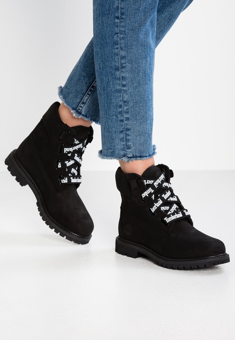 Pieds portant des bottes Timberland noires avec des lacets blancs portant le logo et un jean bleu à ourlet effiloché, debout sur un sol blanc.