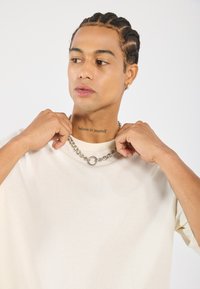 Beige kortermet t-skjorte med rund hals, kombinert med en tykk sølvkjede med en sirkulær anheng.