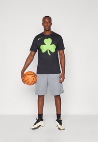 Camiseta negra con un gran diseño de trébol verde, combinada con pantalones cortos grises y zapatillas deportivas en blanco y negro, sosteniendo un baloncesto.