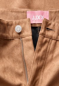 Vêtement en daim marron avec fermeture éclair argentée et bouton, montrant une étiquette rose avec texte blanc "JJXX" à l'intérieur de la ceinture.