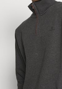 Tmavě šedý pletený svetr s výstřihem do stojáčku, polo zipem a decentním logem na hrudi. Měkká textilie s žebrovanými detaily.