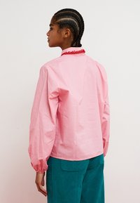 Vaaleanpunainen, kevyt pusero puffyillä hihoilla ja punaisella koristekauluksella, yhdistetty teelimaisiin housuihin. Sileä pinta, rento muotoilu.