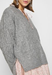 Grauer Strickpullover mit Kabelmuster, V-Ausschnitt und pinkem Band am Hals, kombiniert mit einem pinken gemusterten Kleid.